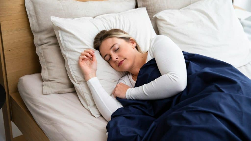 A fair-haired woman is sleeping peacefully under a dark blue blanket on a bed.