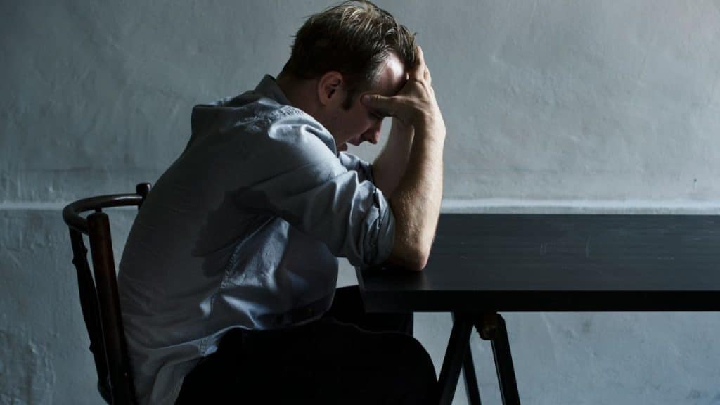 Man seated at a dark table, leaning forward with his hands covering his head.