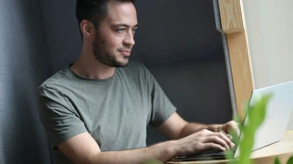Young man with a beard sitting in a wooden cubicle, working on a laptop.