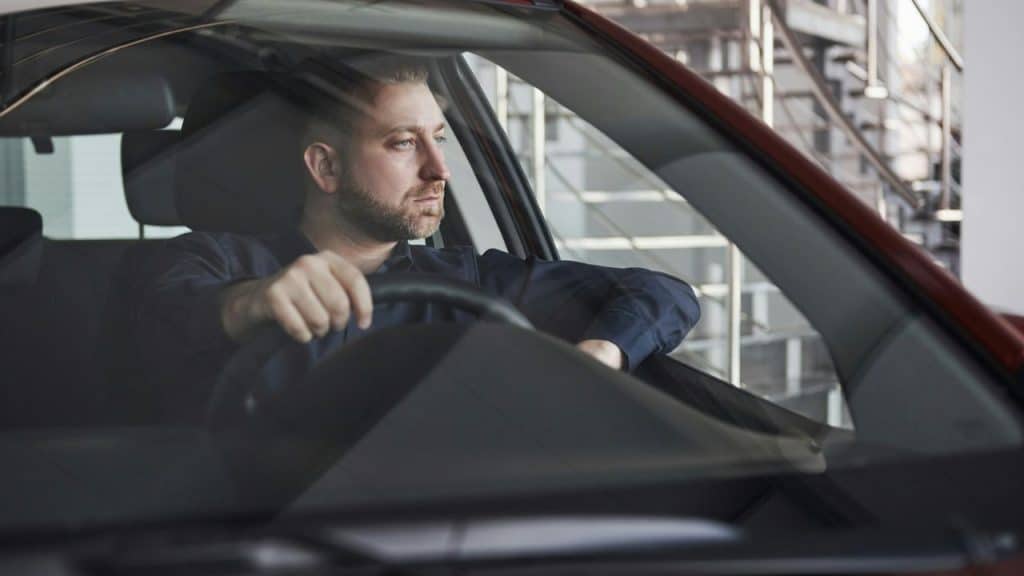 Man with a beard sits in the driver's seat of a car, looking out the window.
