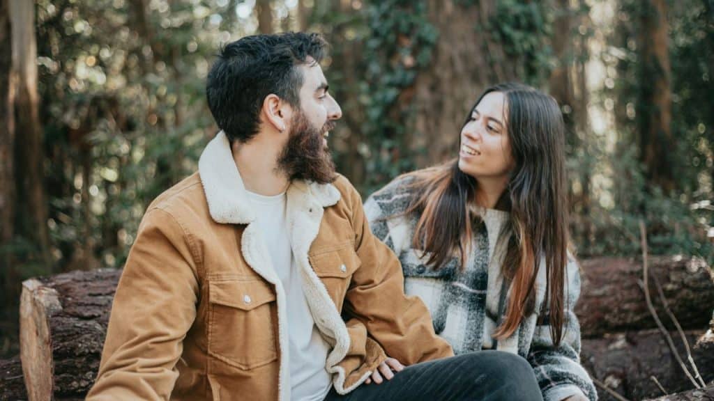 A smiling couple sits on a log in a forest, talking and looking at each other.