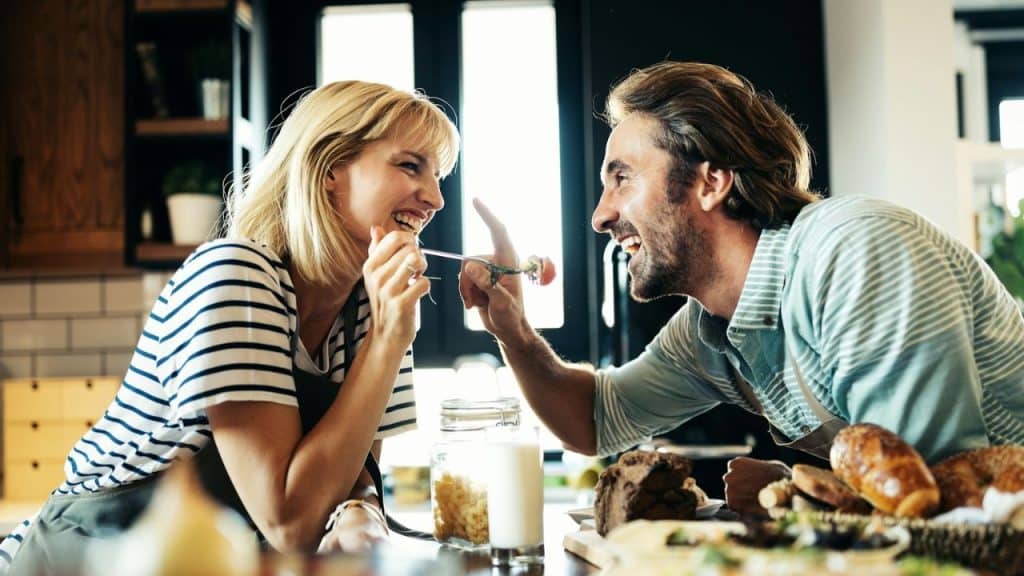 A smiling man in a kitchen is playing with a smiling blonde woman.