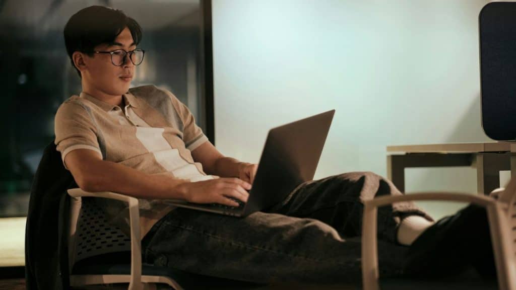 A young man wearing glasses sits in a chair with his feet up, working on a laptop.