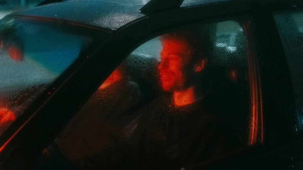 A driver in a car at night, seen through a rainy window with an orange glow on his face.