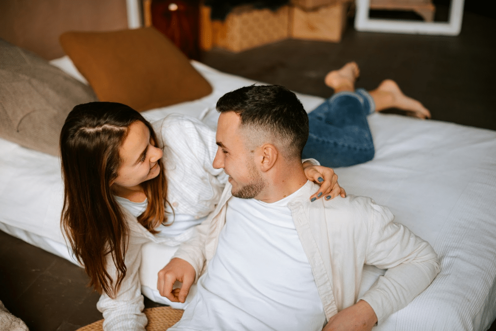 The woman is lying on the bed while the man is sitting on the bedroom floor.