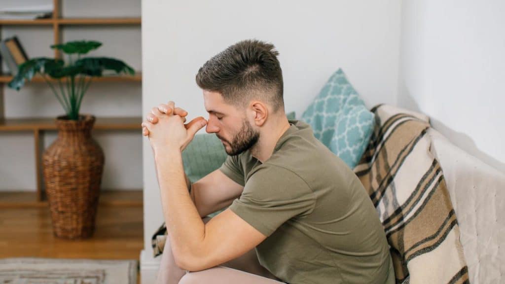 A man sitting on a couch leaning forward with his hands clasped in thought.