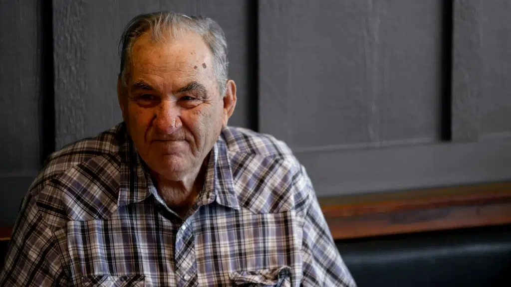 An older man in a plaid shirt sits smiling against a dark, wooden-paneled background.