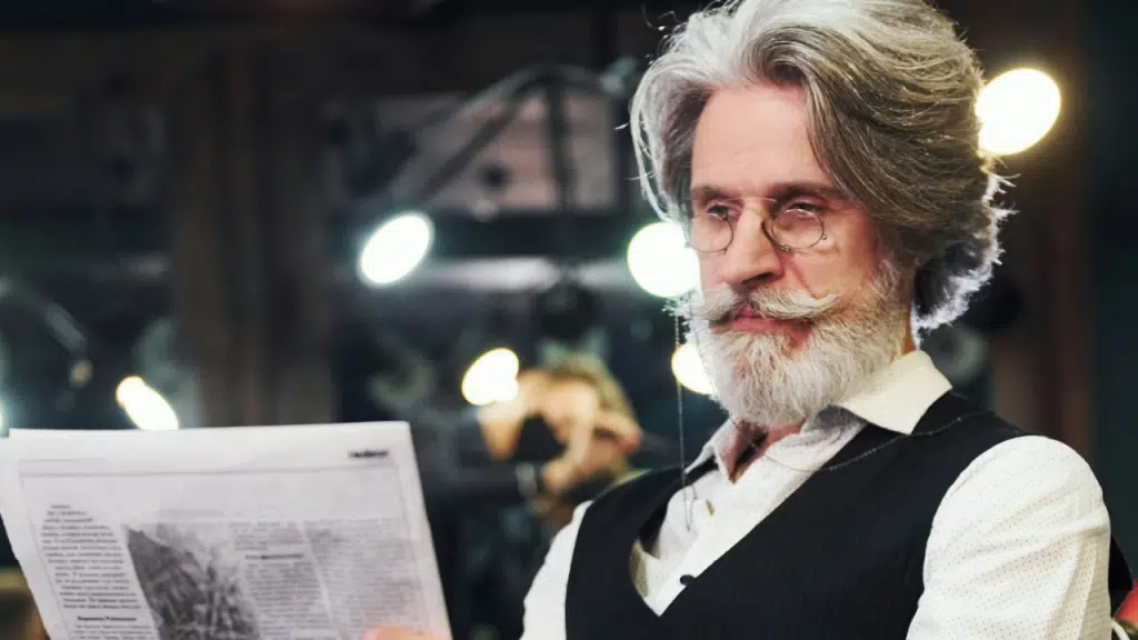 A stylish older man with a long beard and glasses reads a newspaper indoors.