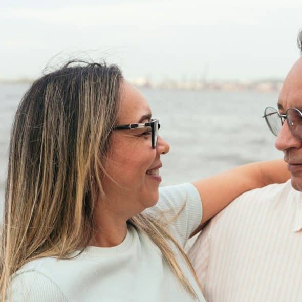 A happy, middle-aged couple, wearing glasses, stands embracing outdoors near the water, smiling warmly at each other.