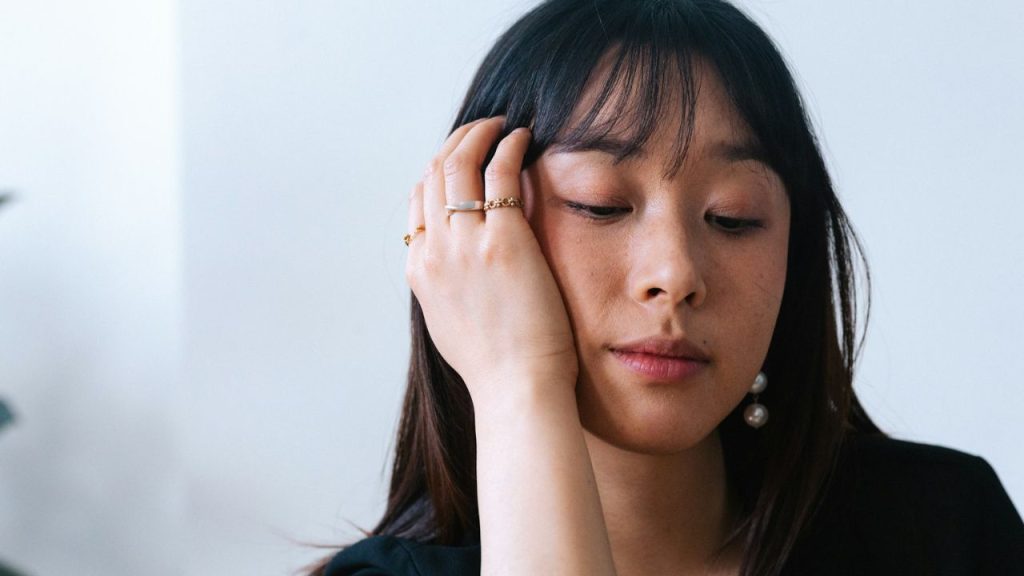 A close-up of a contemplative woman with long dark hair, holding her face with one hand.