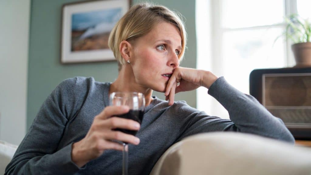 A worried-looking woman with short blonde hair sits on a couch, holding a glass of red wine.