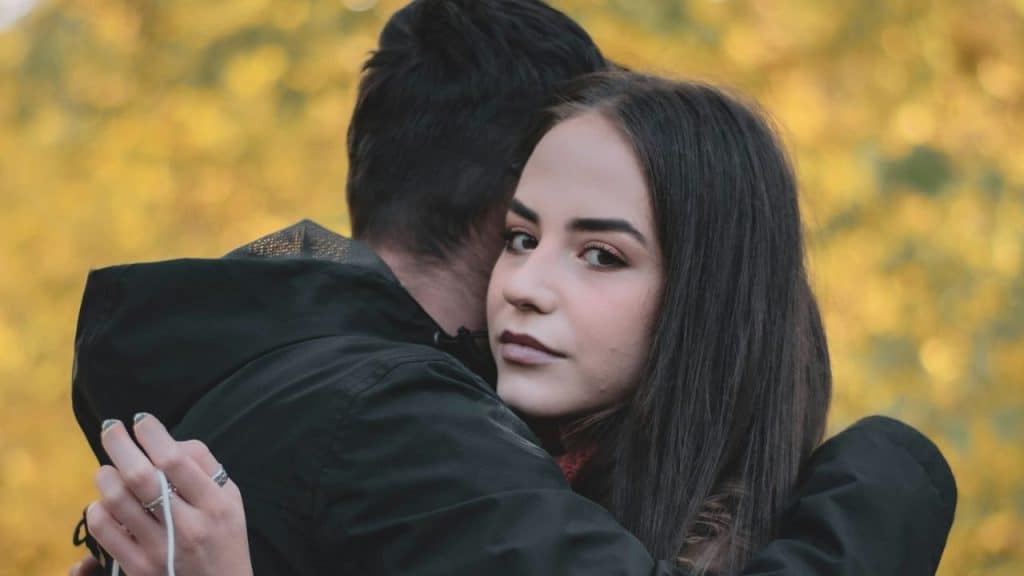 A young woman is hugging a man, looking over his shoulder toward the camera.