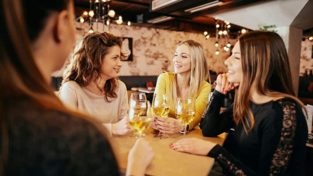 Four women are smiling, sitting at a table with glasses of white wine in a warmly lit restaurant.