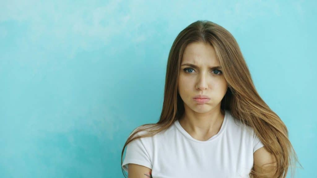 A frustrated woman with long brown hair, wearing a white shirt, is looking at the camera.