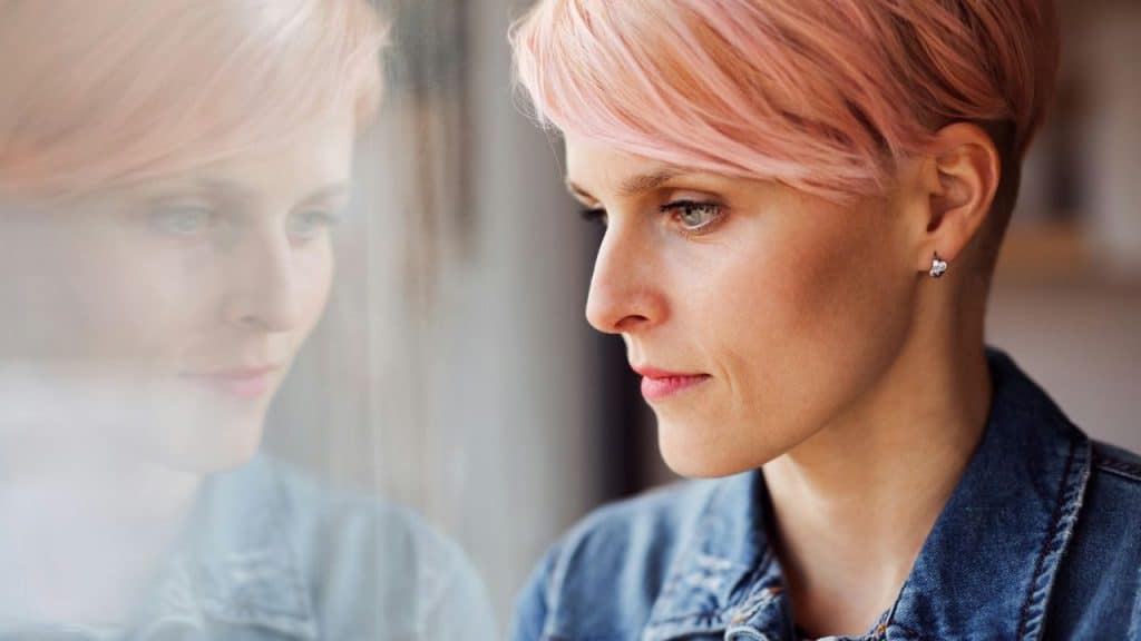 A woman with pink, short hair looks out a window, reflecting her image in the glass.