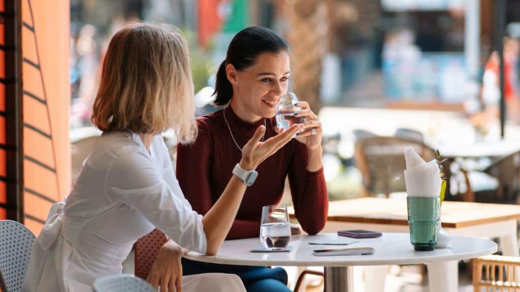 Two women are sitting at an outdoor table, one is drinking water and smiling while talking.