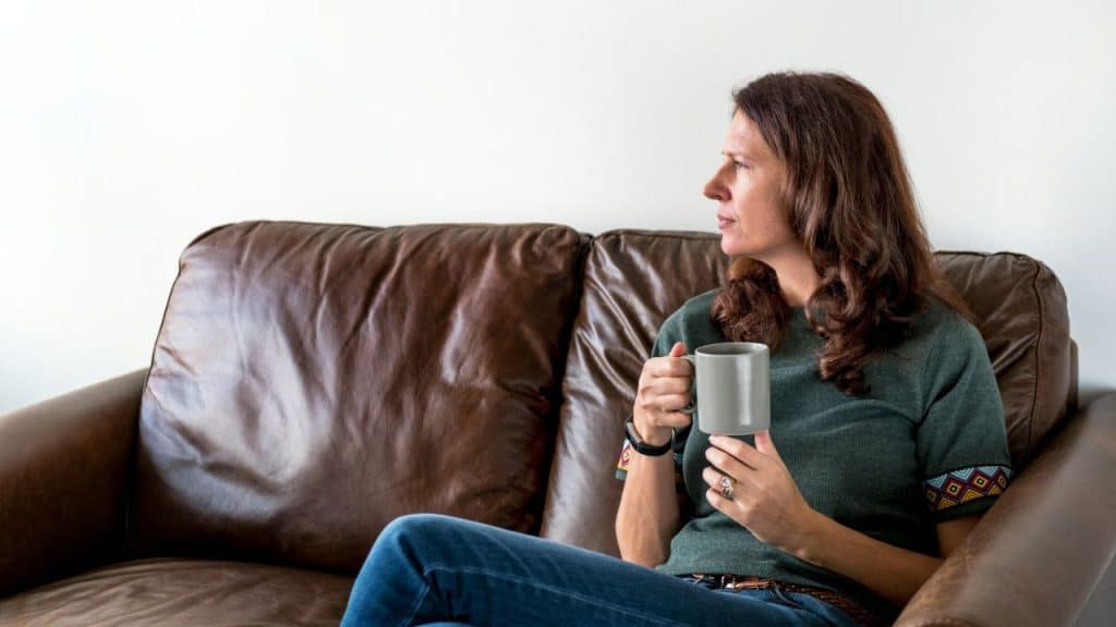 A woman with long brown hair sits on a brown leather couch holding a gray mug.
