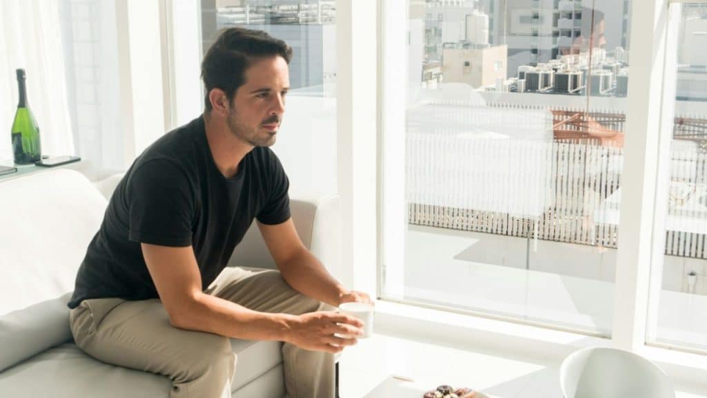 A man in a black t-shirt and khaki pants sits on a white couch looking out a large window.