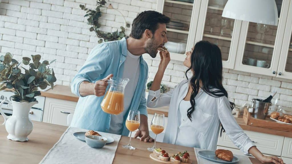 A woman feeds a piece of food to a man holding a pitcher of orange juice in a kitchen.