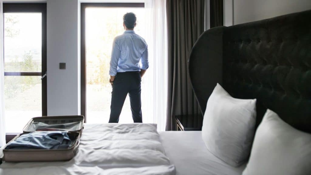 A man in a blue shirt and dark pants stands looking out a bright glass door in a room.