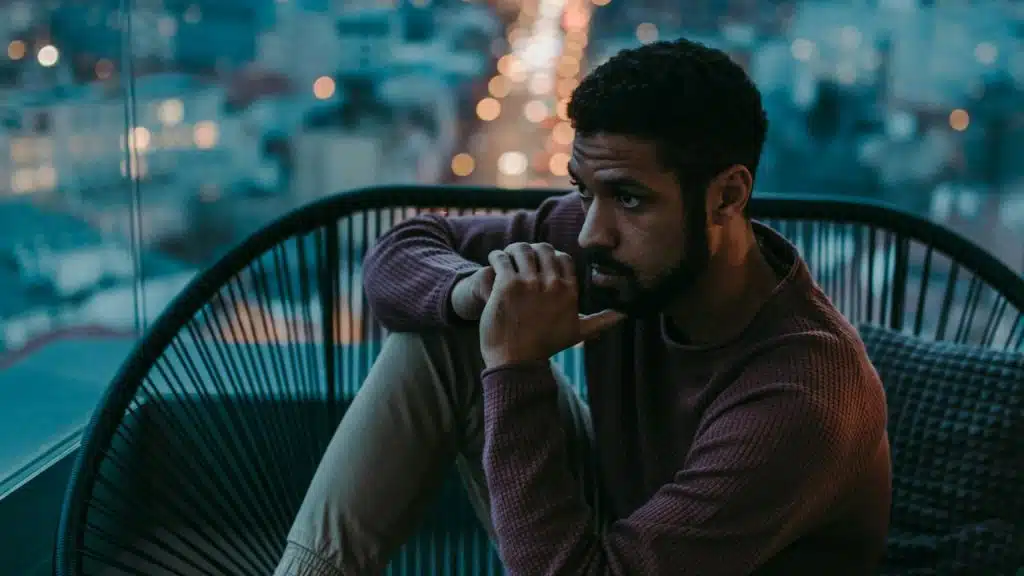 A thoughtful man sits in a black wire chair looking out at a city skyline at night.