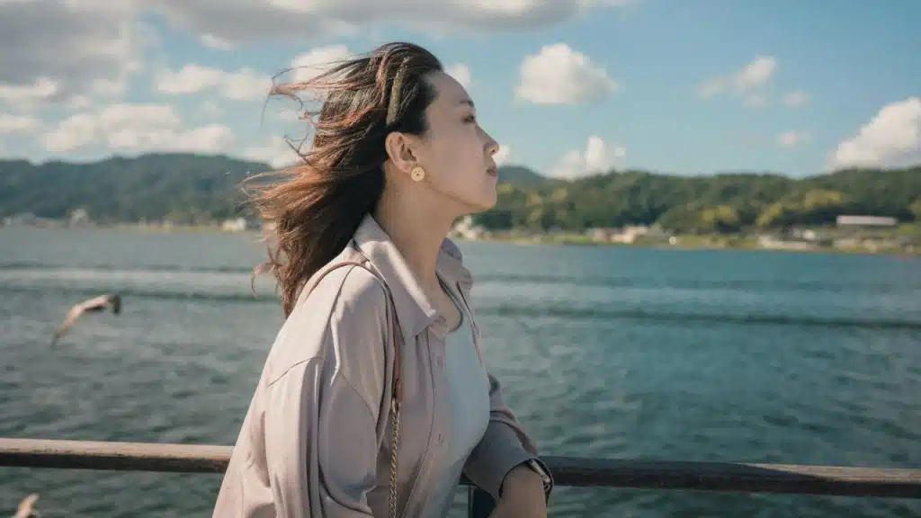 A woman with wind-blown hair stands by a lake or sea, looking out under a blue sky.