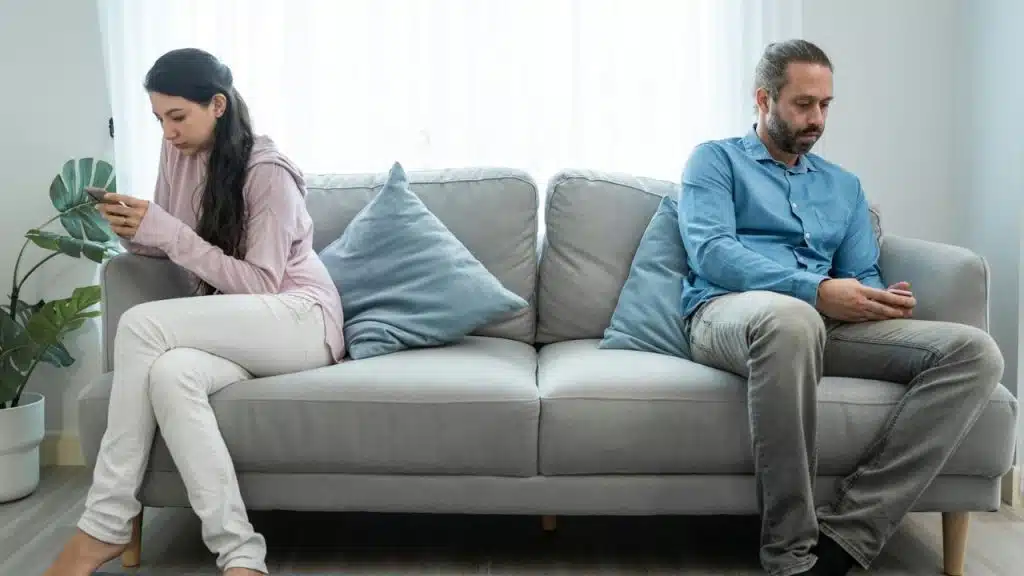 A man and woman sit far apart on a couch, both looking down at their mobile phones.