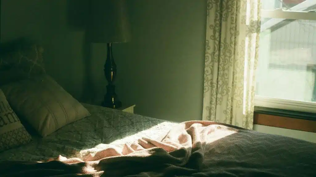 Sunlight streams onto an unmade bed next to a window and a lamp in a dimly lit room.