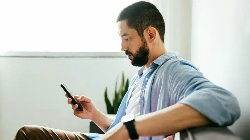 A bearded man in a light blue shirt sits on a sofa, looking intently at a mobile phone.