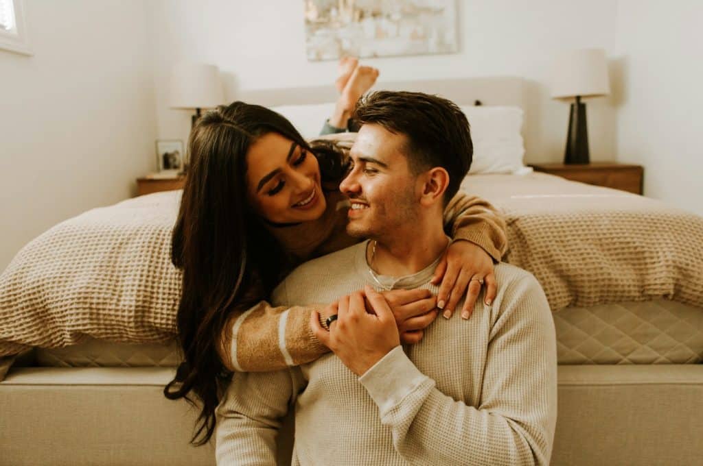The woman is hugging the man who’s sitting on the bedroom floor.