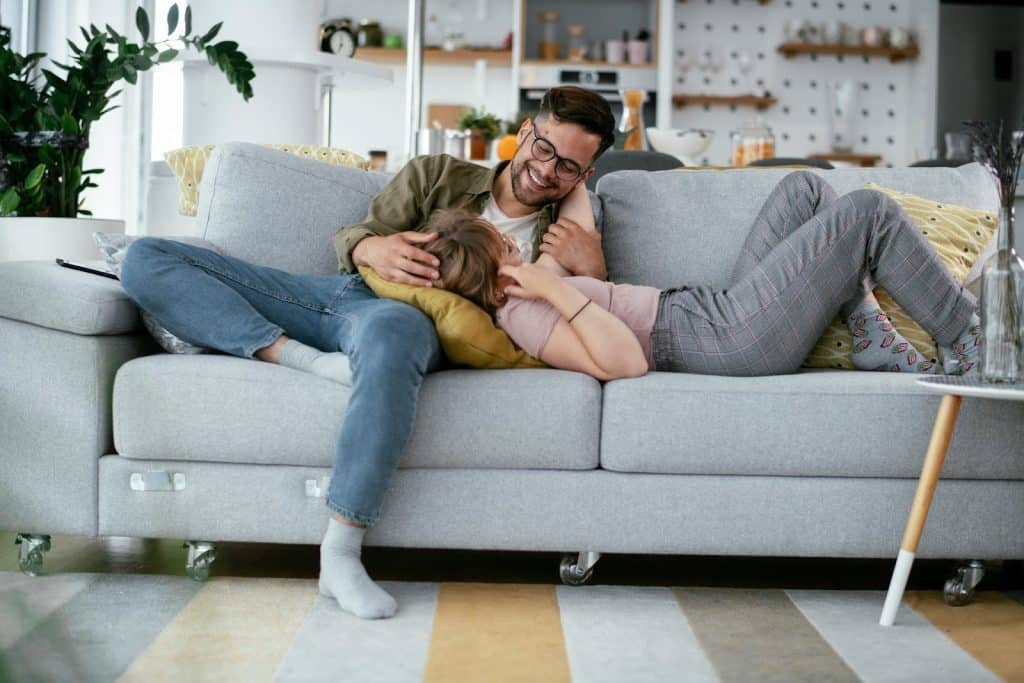 The husband and wife are lying on the gray couch in the living room.