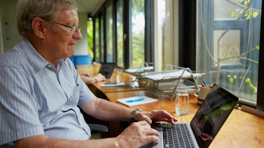 An older man with glasses works on a laptop at a wooden desk with large windows.