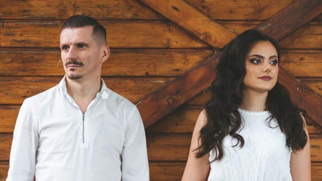 A stern-looking couple stands next to each other in front of a wooden background.