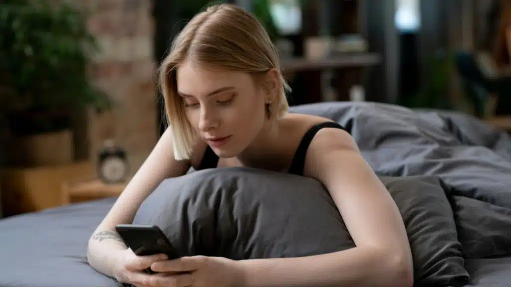 A blonde woman is lying in bed, leaning on a pillow and looking at her smartphone.