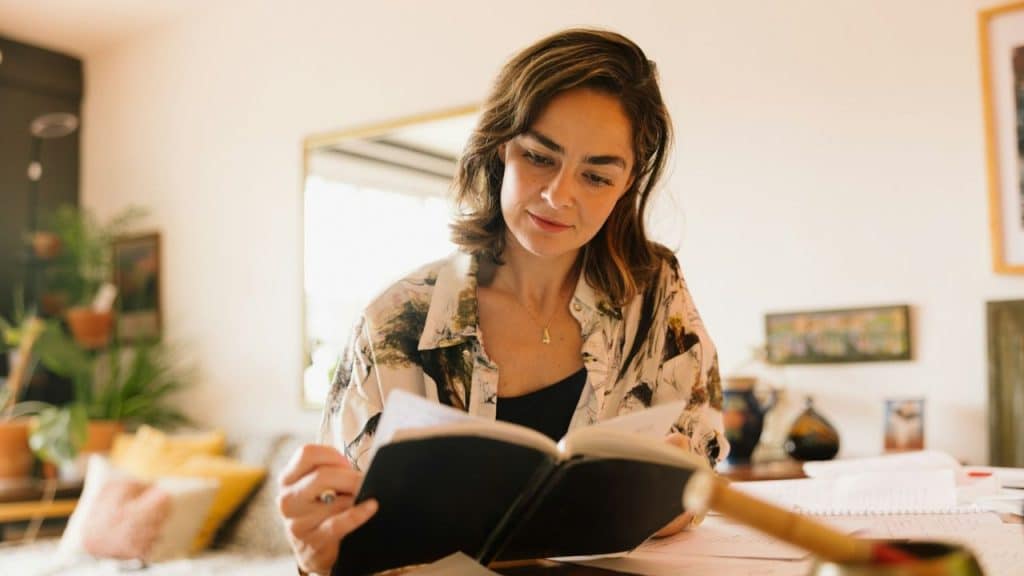 A focused woman in a patterned shirt is sitting indoors, reading a large black notebook.