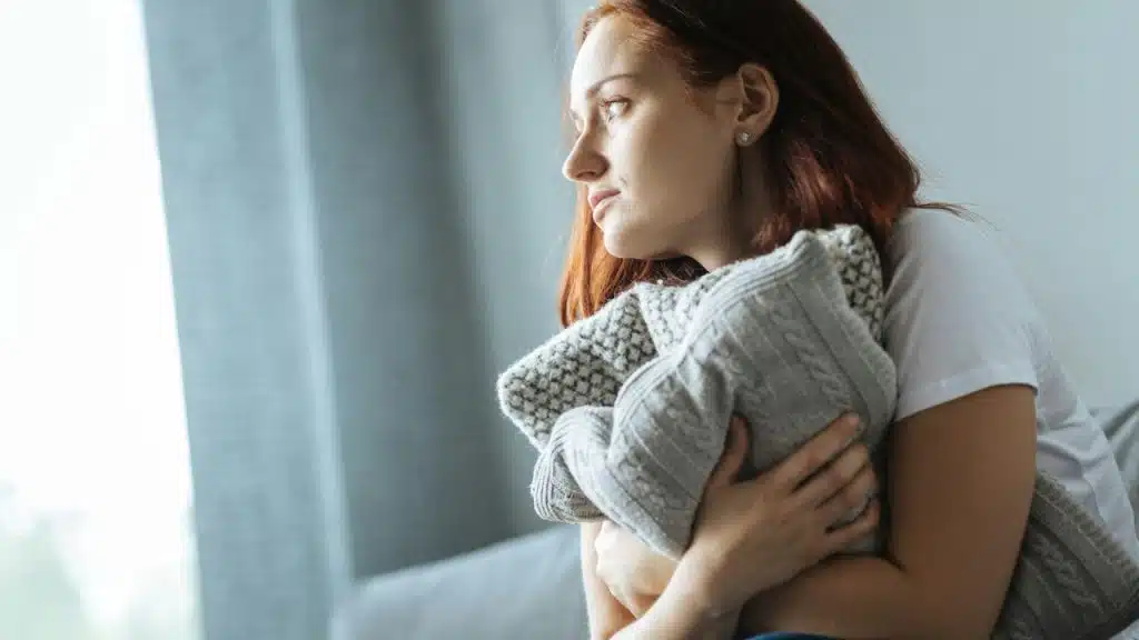A woman with auburn hair is hugging a gray knitted pillow while looking out a window.