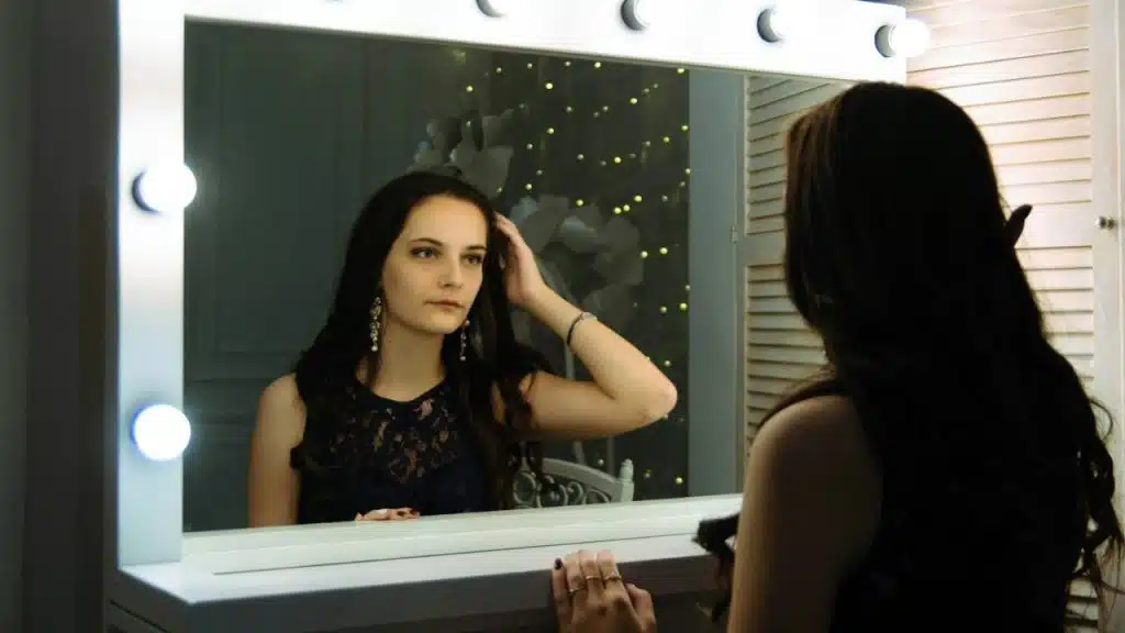 A woman in a dark lace top is looking at her reflection in a lighted vanity mirror.