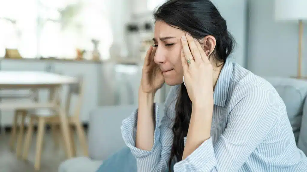 A distressed woman is sitting on a couch, holding her temples with both hands.