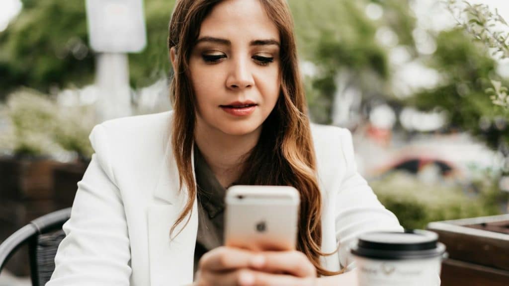 A focused woman in a white blazer is sitting outdoors, looking intently at her smartphone.