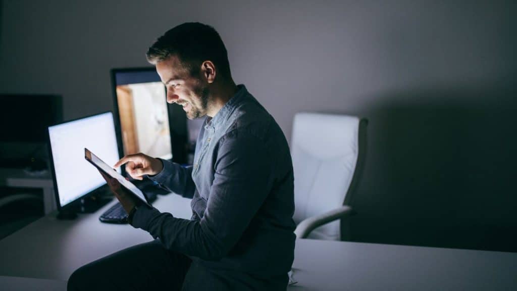 A smiling man sits on a desk at night, working on a tablet in an office.