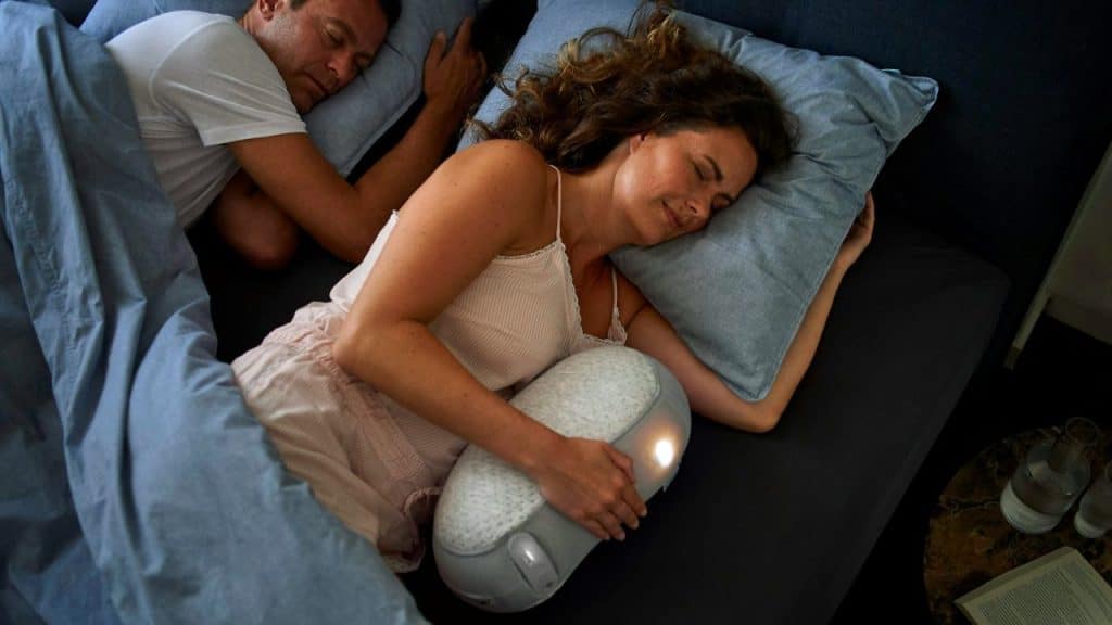 A man and woman are asleep in bed; the woman is hugging a glowing, oval-shaped object.