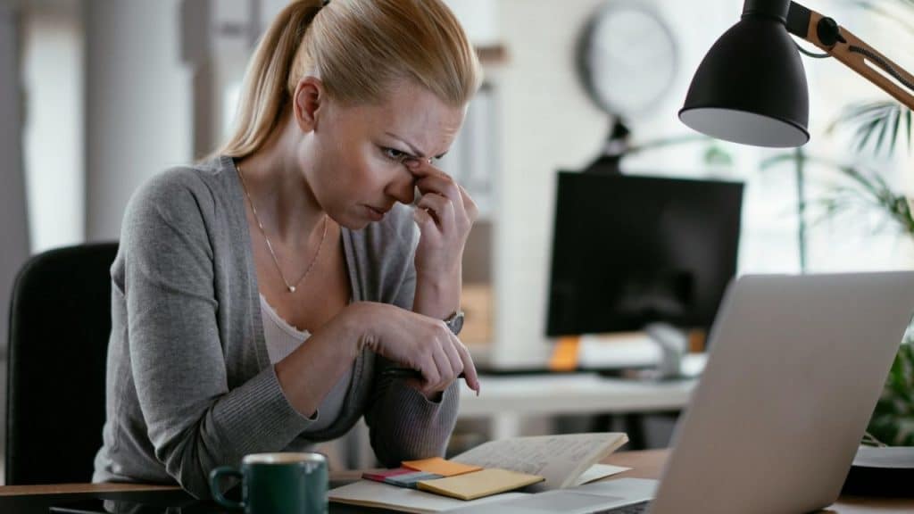 A distressed woman is sitting at a desk, rubbing the bridge of her nose while looking at a laptop.