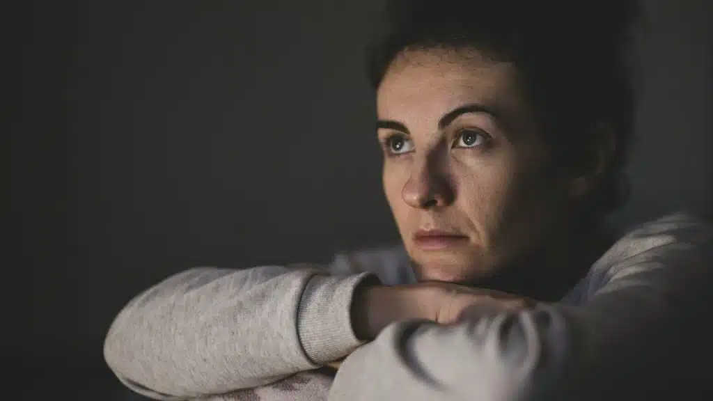 A serious woman is leaning on her forearms, looking up and away from the camera in low light.