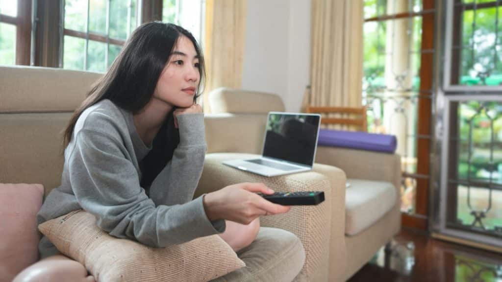 A young woman sits on a couch, holding a remote control and watching television.