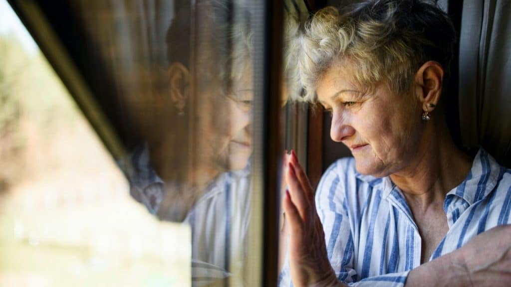 An older woman with short gray hair is looking out a window with her hand resting on the glass.