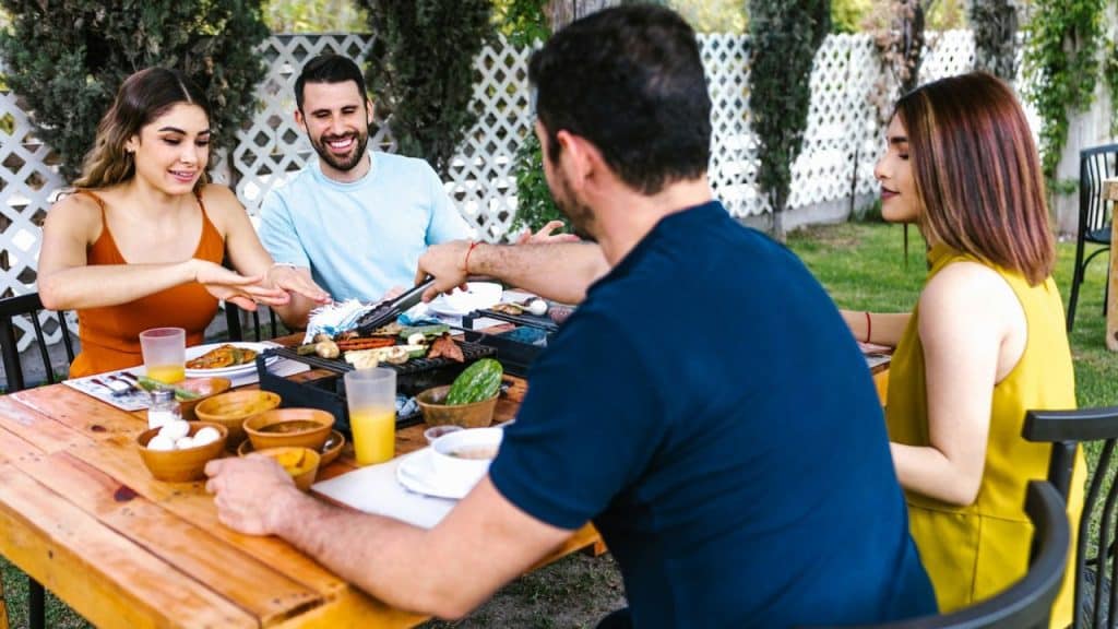 Four people are gathered around an outdoor wooden table, enjoying a barbecue.