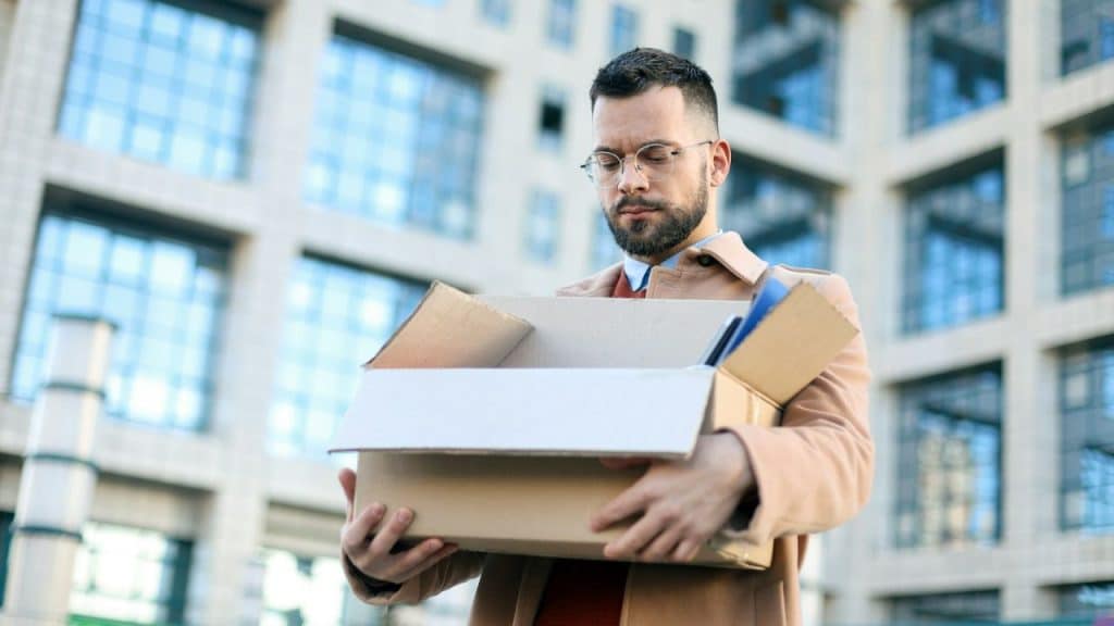 A serious man in a trench coat and glasses is standing outdoors, holding a box of personal items.