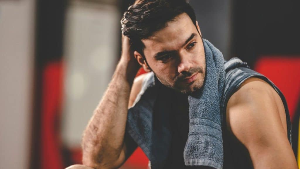 A pensive, sweaty man with a towel around his neck is sitting in a gym.