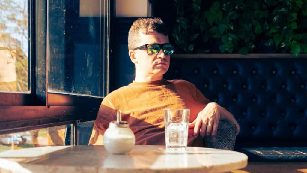 A serious man wearing mirrored sunglasses sits at a table in a dimly lit cafe.