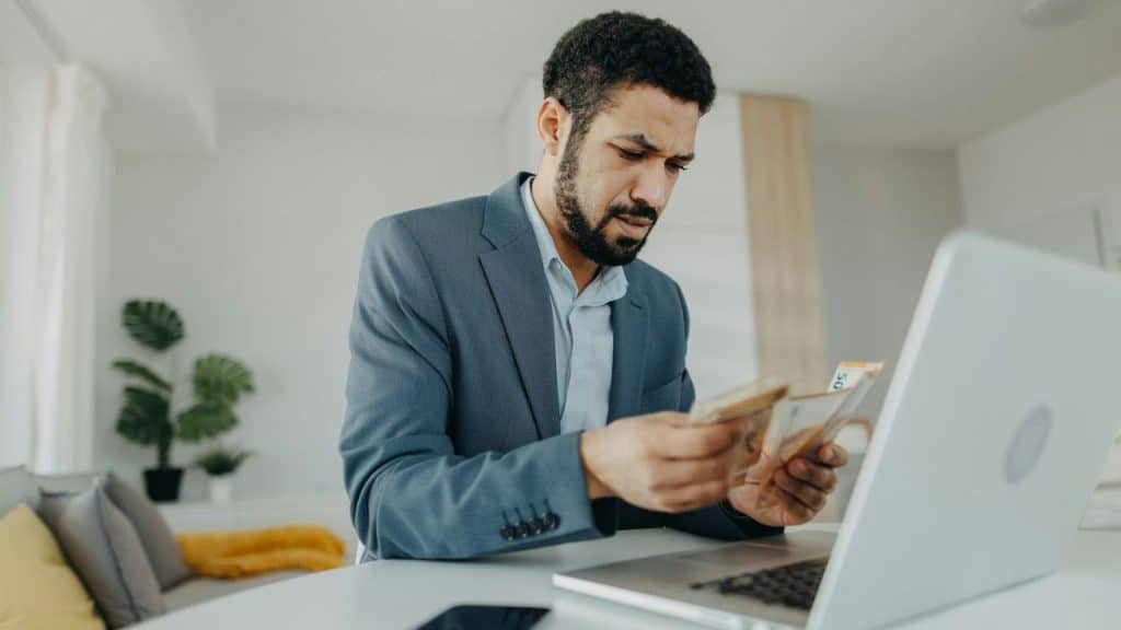A bearded man in a suit is sitting at a laptop, intently counting banknotes.
